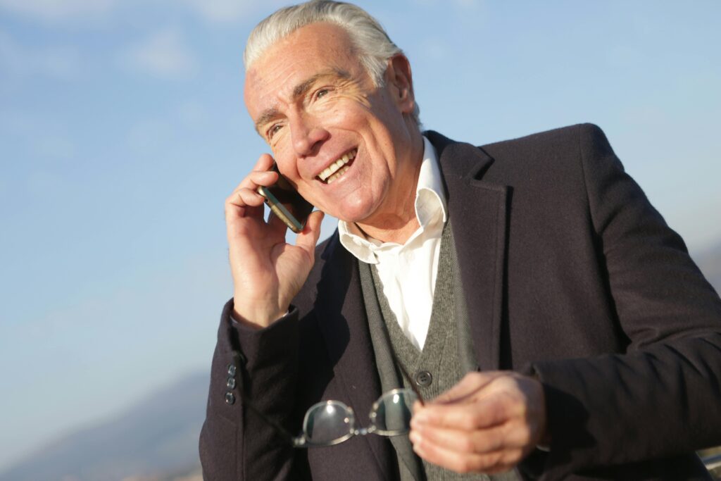 Joyful senior man in formal attire speaking on a cellphone while enjoying a sunny day outdoors.